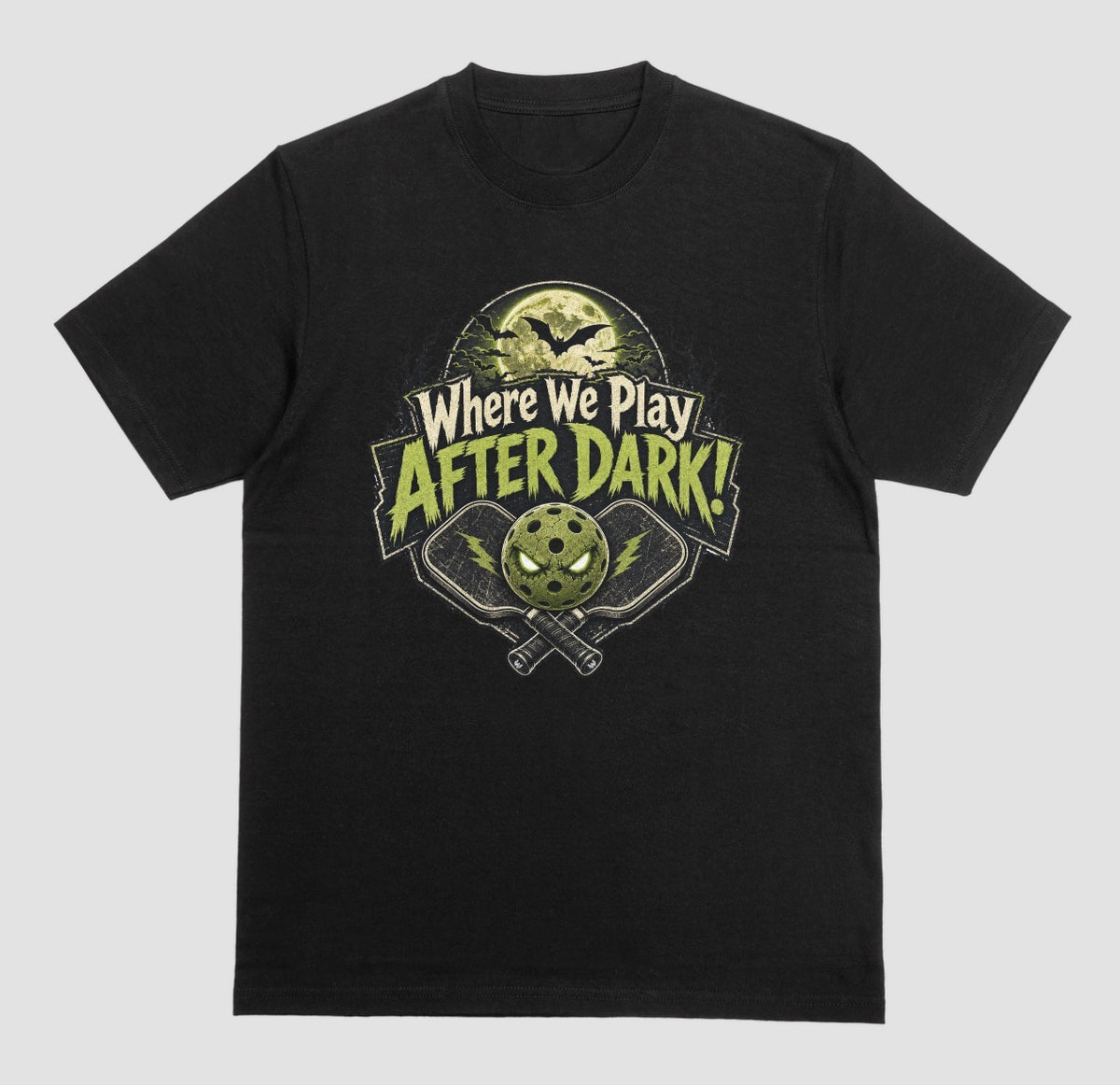 A black t-shirt with a spooky pickleball graphic reading 'Where We Play After Dark!' showing crossed paddles and a glowing-eyed pickleball under a full moon with bats.