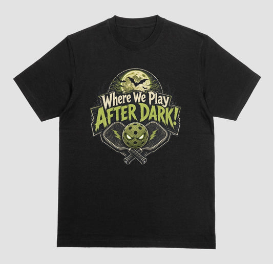 A black t-shirt with a spooky pickleball graphic reading 'Where We Play After Dark!' showing crossed paddles and a glowing-eyed pickleball under a full moon with bats.