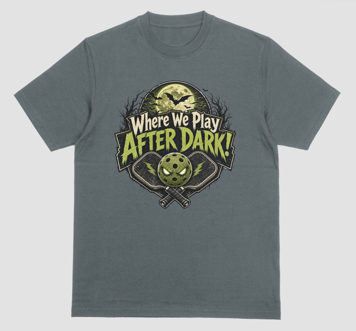 A dark heather t-shirt with a spooky pickleball graphic reading 'Where We Play After Dark!' showing crossed paddles and a glowing-eyed pickleball under a full moon with bats.