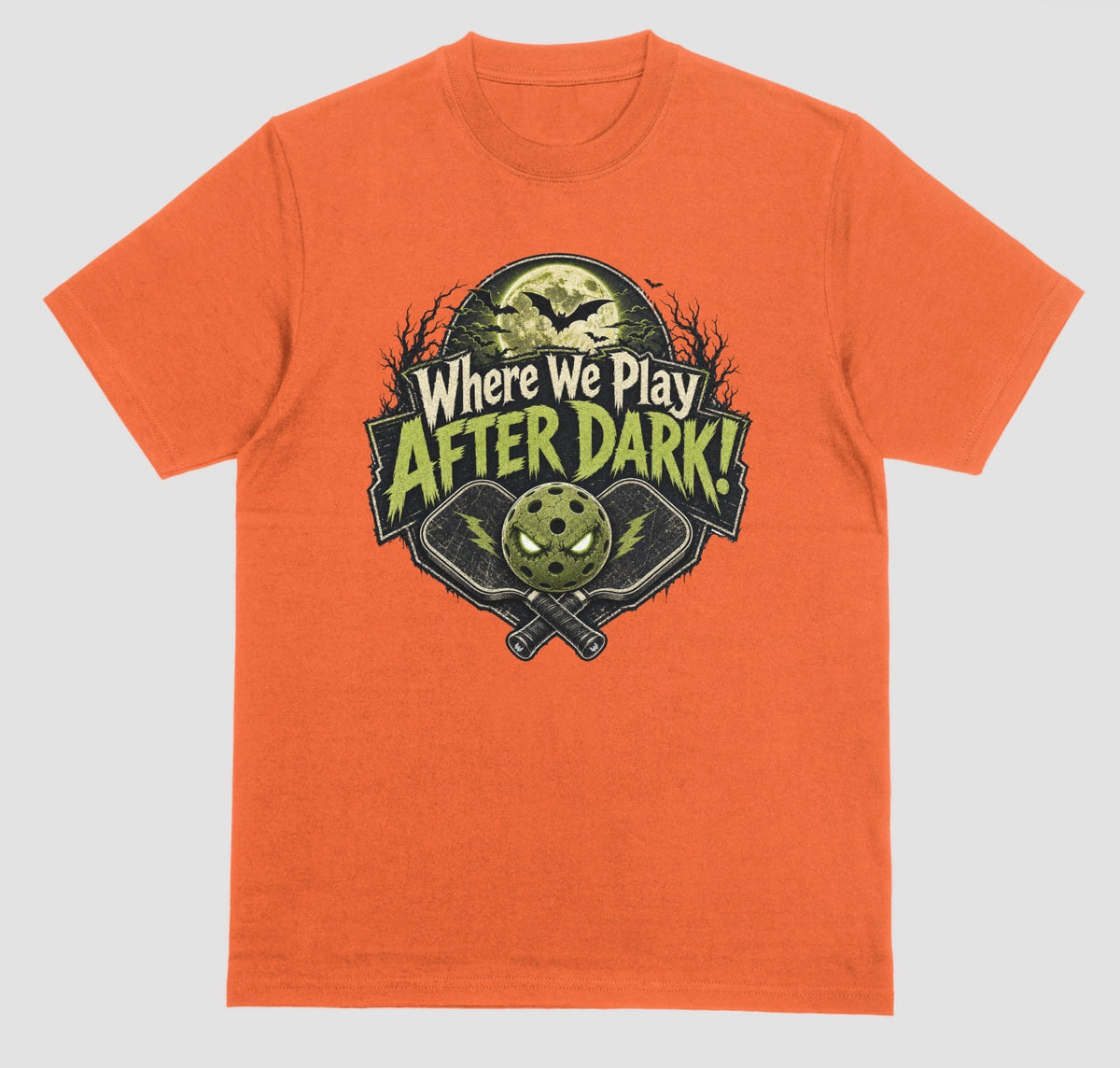An orange t-shirt with a spooky pickleball graphic reading 'Where We Play After Dark!' showing crossed paddles and a glowing-eyed pickleball under a full moon with bats.