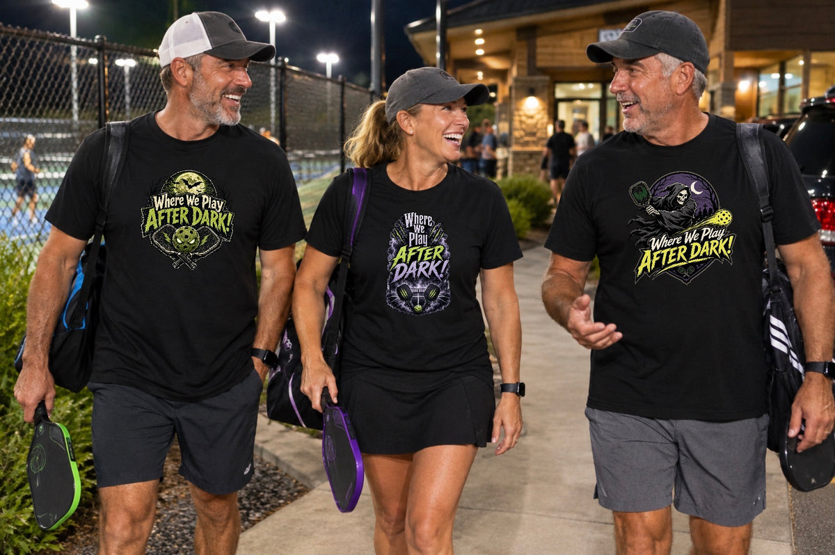 "Two men and a woman walk along a sidewalk beside a lit pickleball court at night, smiling and chatting with paddles and gear bags in hand. All three wear black 'Where We Play After Dark' t-shirts with different haunted pickleball graphics: a lime-green design with crossed paddles and a glowing-eyed ball, a purple-and-green design with a lightning-streaked storm cloud, and a yellow-and-purple design with a hooded grim reaper swinging a paddle under a crescent moon."