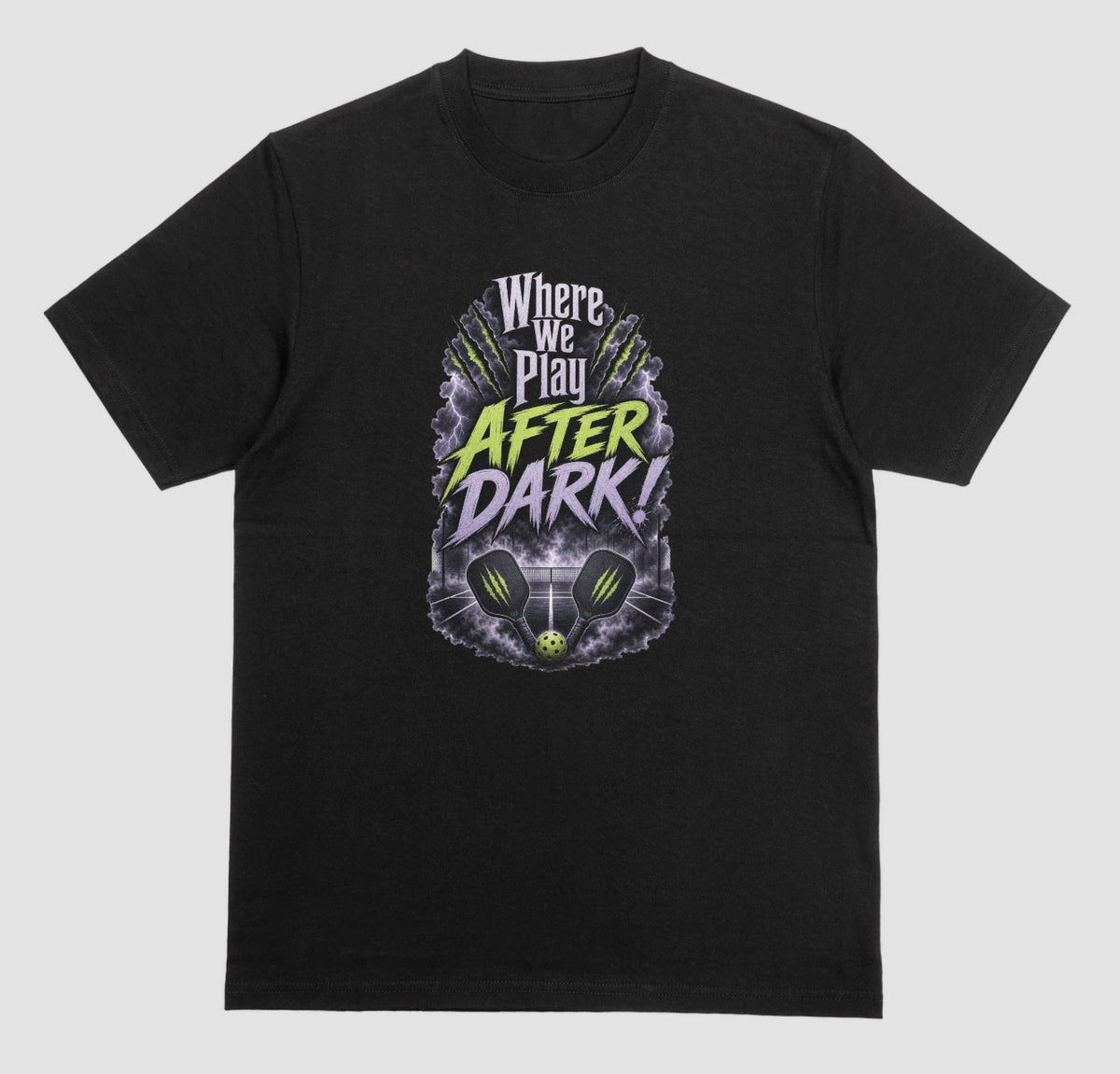 A black t-shirt with a spooky pickleball graphic reading 'Where We Play After Dark!' showing two crossed paddles and a ball on a court inside a lightning-filled storm cloud.