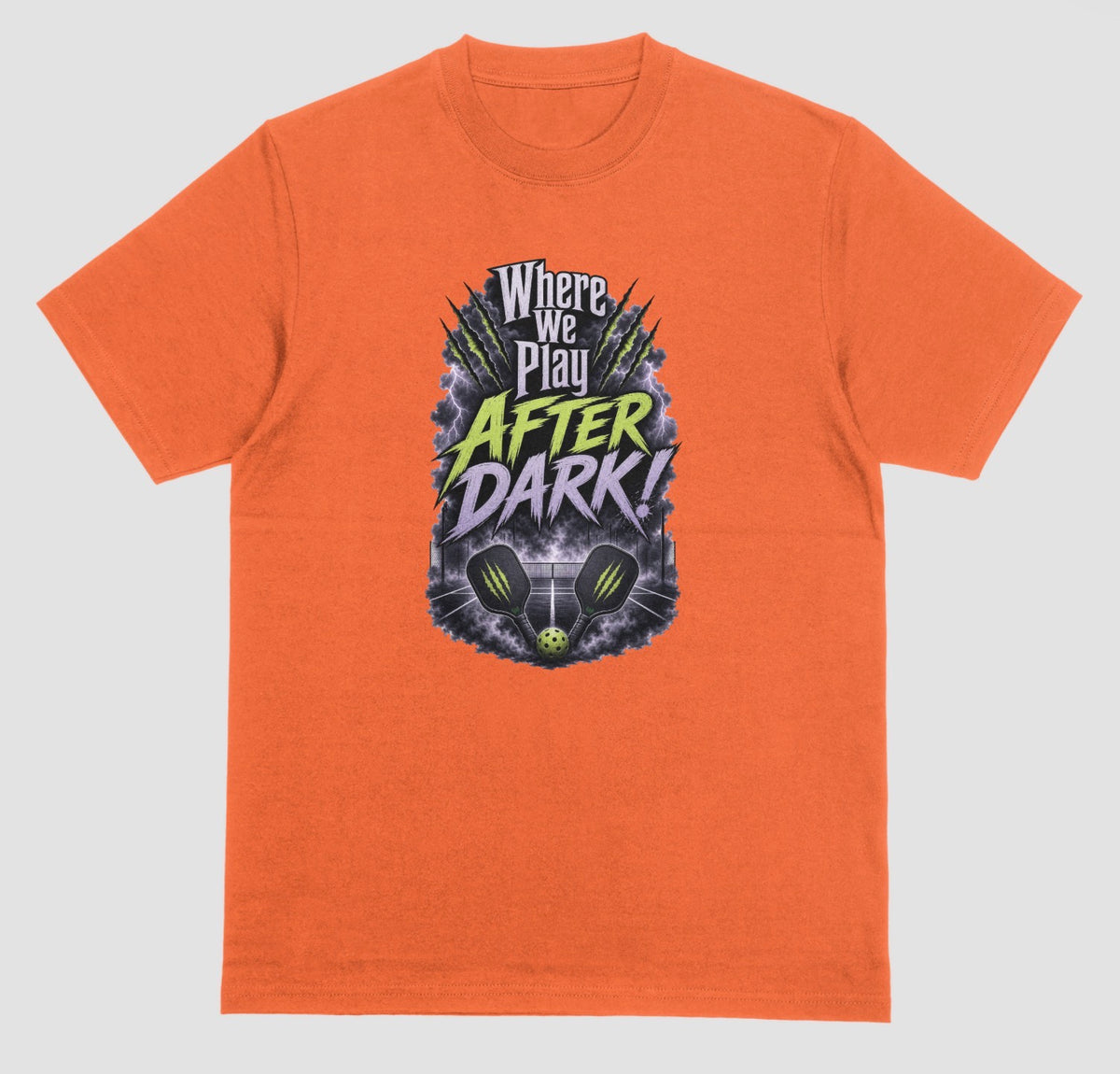 An orange t-shirt with a spooky pickleball graphic reading 'Where We Play After Dark!' showing two crossed paddles and a ball on a court inside a lightning-filled storm cloud.