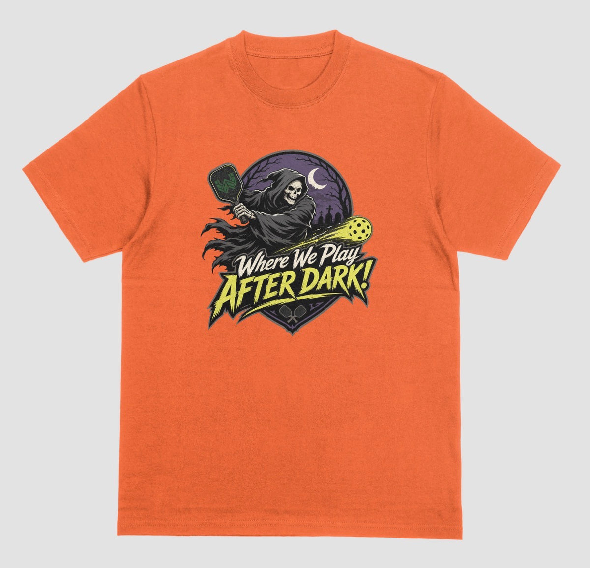 An orange t-shirt with a spooky pickleball graphic reading 'Where We Play After Dark!' showing a hooded grim reaper swinging a paddle at a glowing yellow ball.