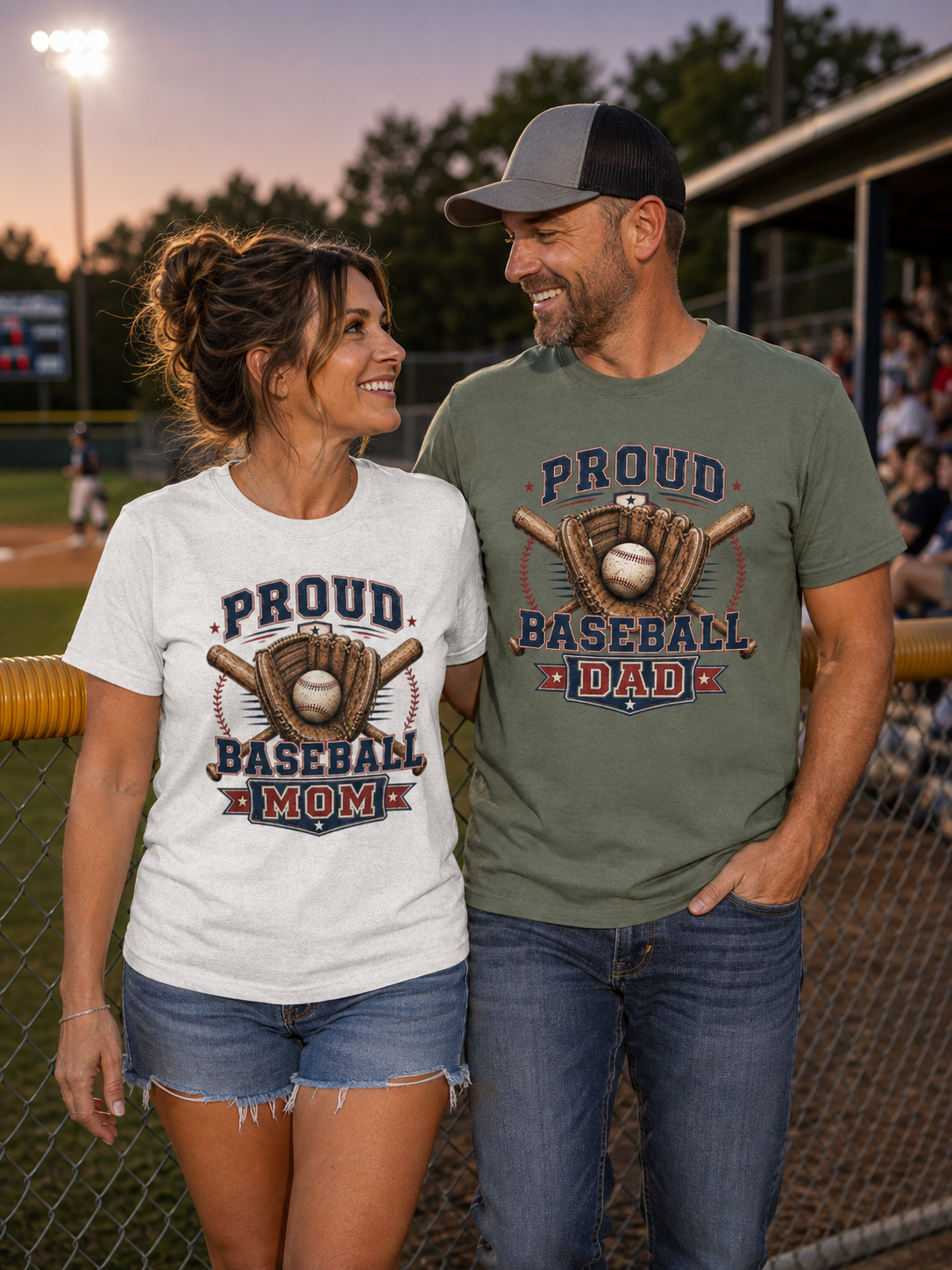 A man and woman standing together at a baseball field at dusk, wearing matching graphic t-shirts. The woman wears a white t-shirt that says 'PROUD BASEBALL MOM' with a baseball glove and bat design, and the man wears a military green t-shirt that says 'PROUD BASEBALL DAD' with the same baseball glove and bat design.