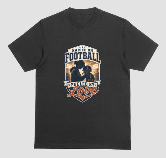 A black crewneck t-shirt with a vintage-style football graphic on the chest. The graphic features a silhouette of a football player with the text 'RAISED ON FOOTBALL' and 'FUELED BY Love' in navy, white, and orange.