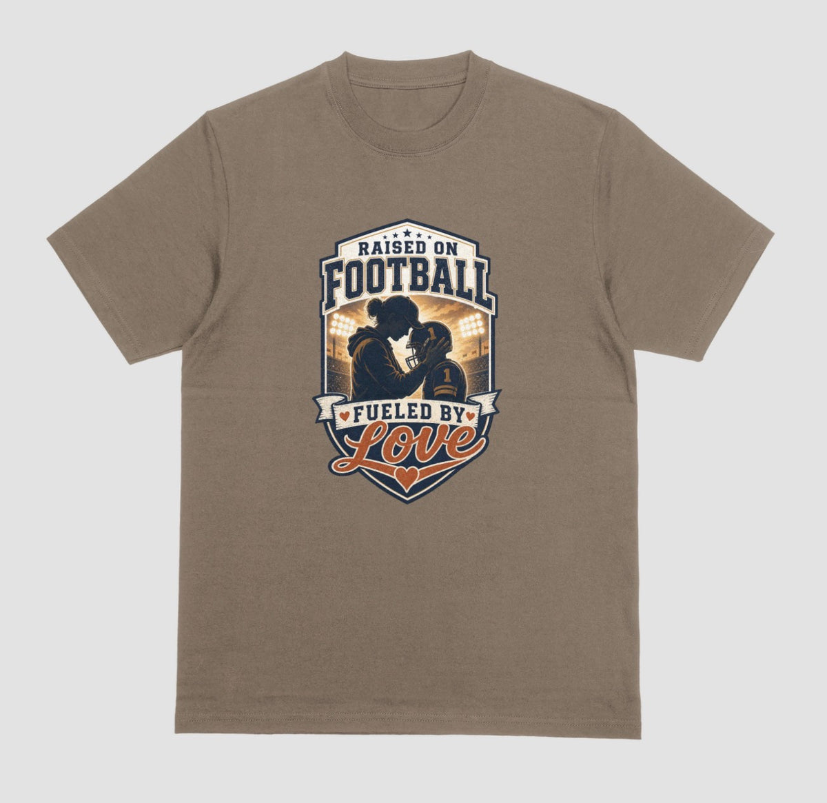 A brown savana crewneck t-shirt with a vintage-style football graphic on the chest. The graphic features a silhouette of a football player with the text 'RAISED ON FOOTBALL' and 'FUELED BY Love' in navy, white, and orange.
