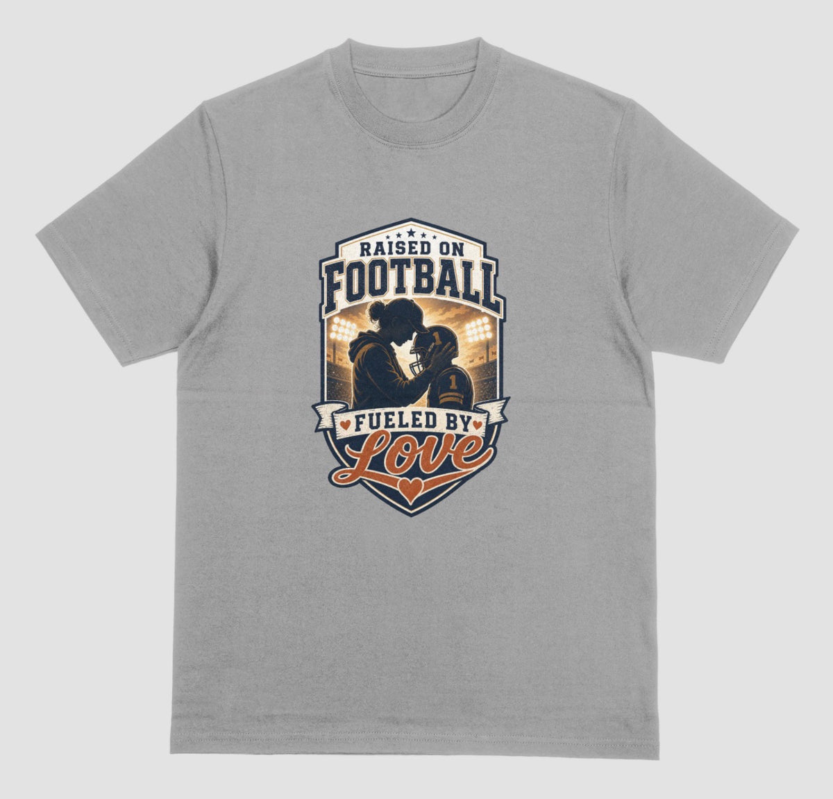 A grey crewneck t-shirt with a vintage-style football graphic on the chest. The graphic features a silhouette of a football player with the text 'RAISED ON FOOTBALL' and 'FUELED BY Love' in navy, white, and orange.