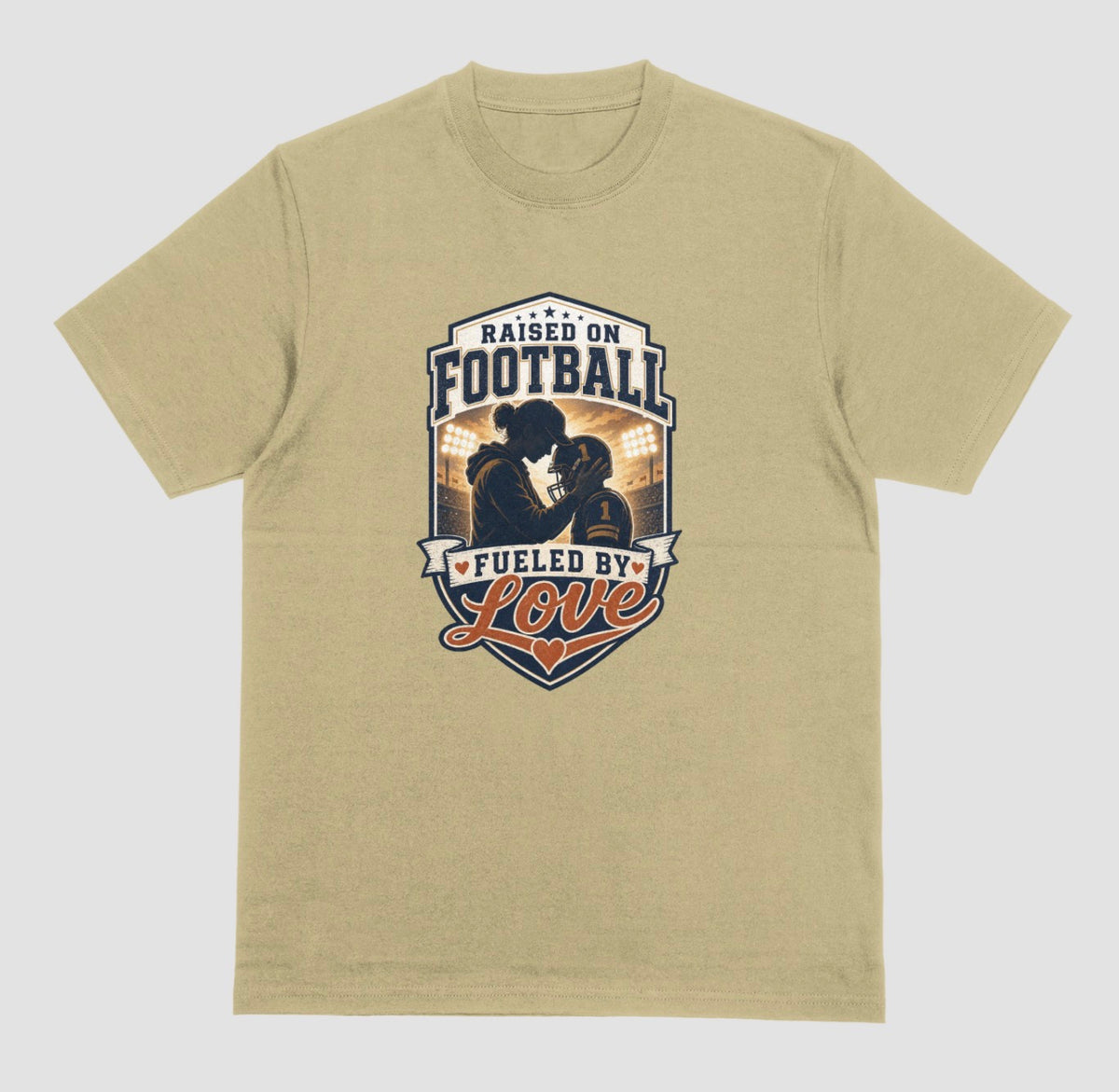 A sand color crewneck t-shirt with a vintage-style football graphic on the chest. The graphic features a silhouette of a football player with the text 'RAISED ON FOOTBALL' and 'FUELED BY Love' in navy, white, and orange.