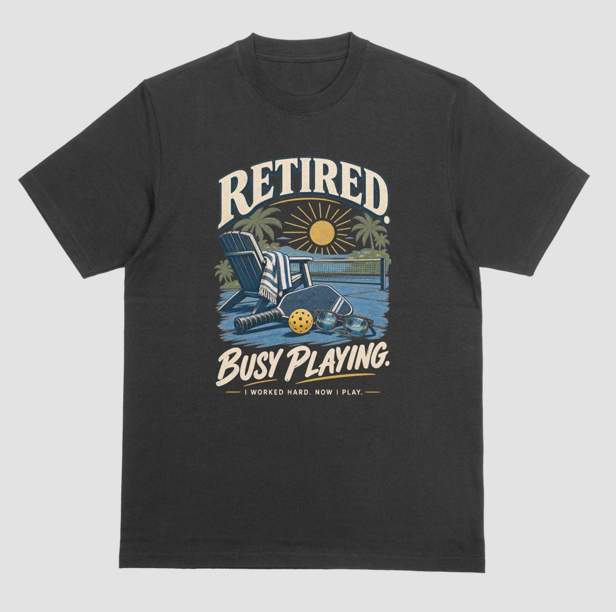 A black short-sleeve t-shirt with a retro graphic on the chest. The graphic features the words short-sleeve t-shirt with a retro graphic on the chest. The graphic features the words Retired pickleball shirt in sand with a beach chair and paddle design that says Retired, Busy Playing, I Worked Hard Now I Play