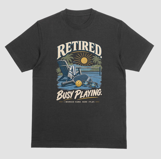 A black short-sleeve t-shirt with a retro graphic on the chest. The graphic features the words short-sleeve t-shirt with a retro graphic on the chest. The graphic features the words Retired pickleball shirt in sand with a beach chair and paddle design that says Retired, Busy Playing, I Worked Hard Now I Play