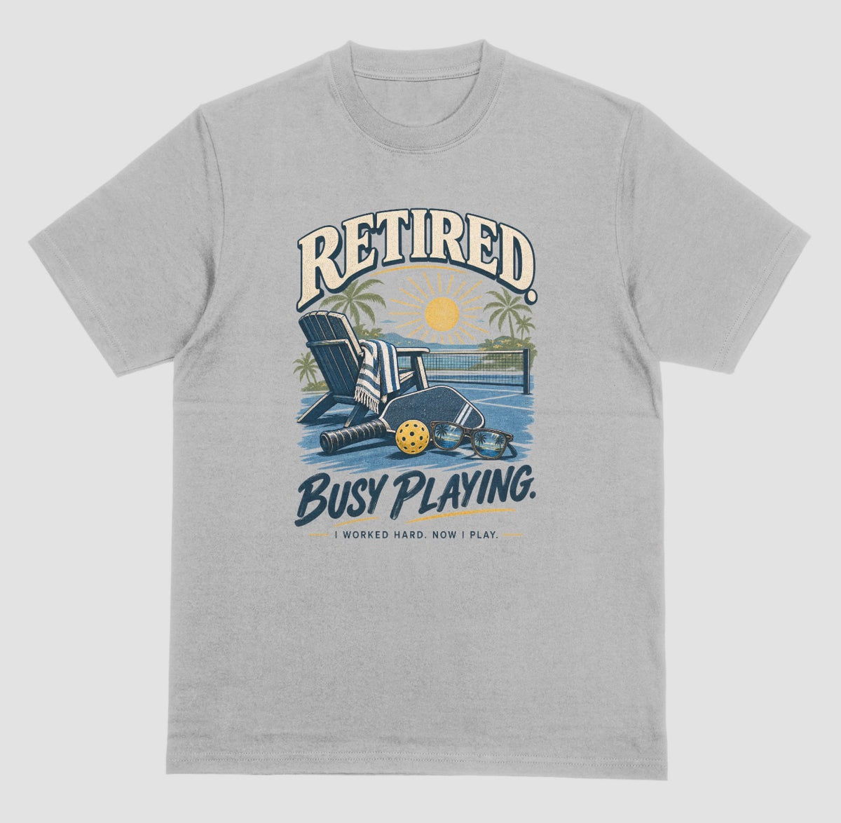 A grey short-sleeve t-shirt with a retro graphic on the chest. The graphic features the words short-sleeve t-shirt with a retro graphic on the chest. The graphic features the words Retired pickleball shirt in sand with a beach chair and paddle design that says Retired, Busy Playing, I Worked Hard Now I Play