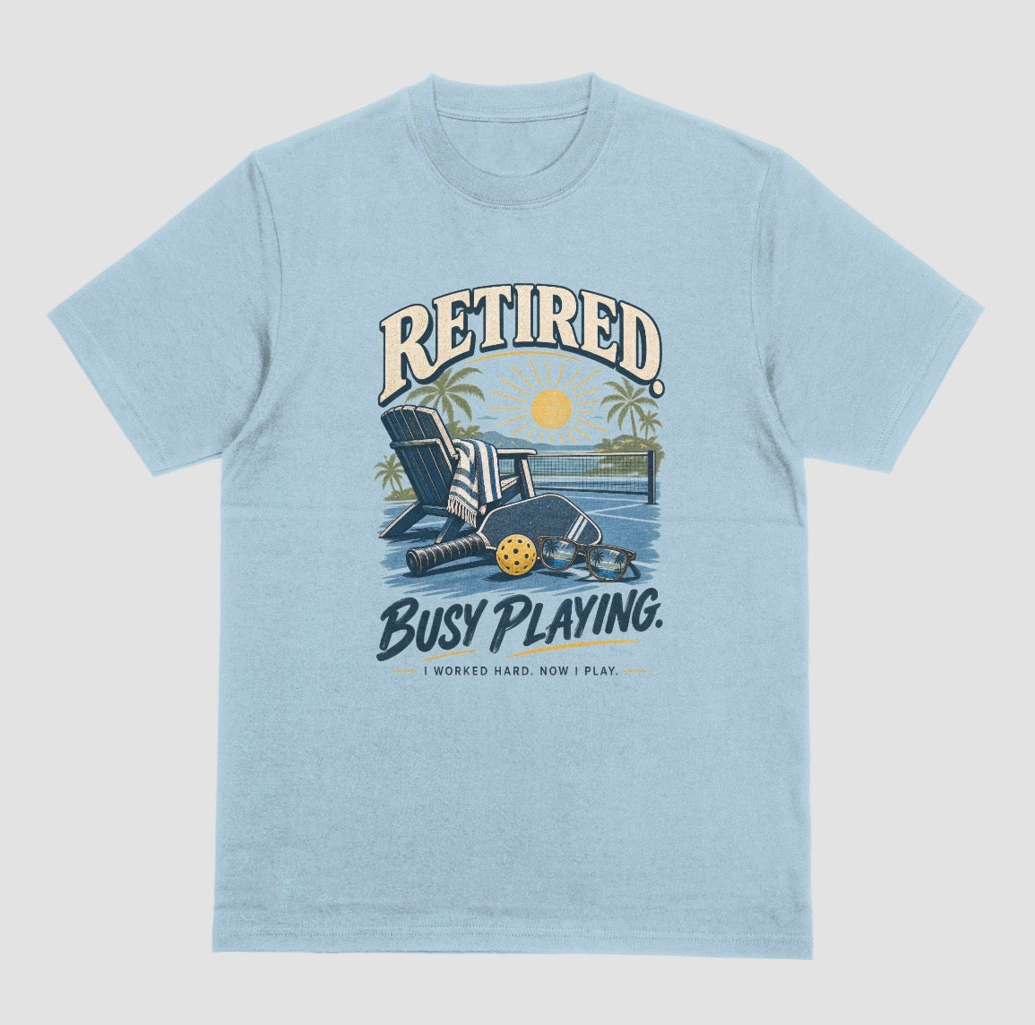 A light blue short-sleeve t-shirt with a retro graphic on the chest. The graphic features the words short-sleeve t-shirt with a retro graphic on the chest. The graphic features the words Retired pickleball shirt in sand with a beach chair and paddle design that says Retired, Busy Playing, I Worked Hard Now I Play