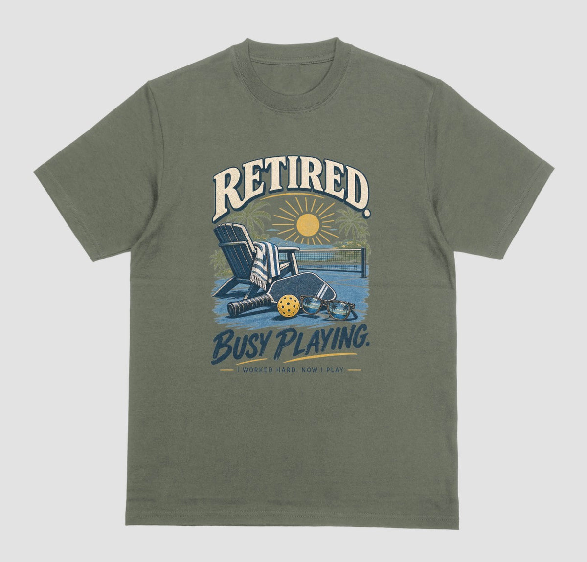 A military green short-sleeve t-shirt with a retro graphic on the chest. The graphic features the words Retired pickleball shirt in sand with a beach chair and paddle design that says Retired, Busy Playing, I Worked Hard Now I Play