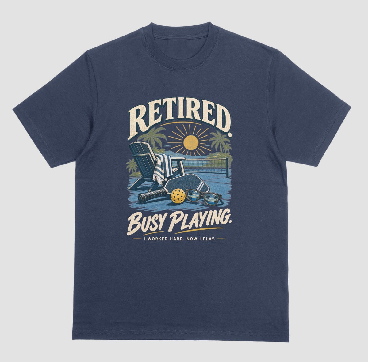 A navy blue short-sleeve t-shirt with a retro graphic on the chest. The graphic features the words Retired pickleball shirt in sand with a beach chair and paddle design that says Retired, Busy Playing, I Worked Hard Now I Play