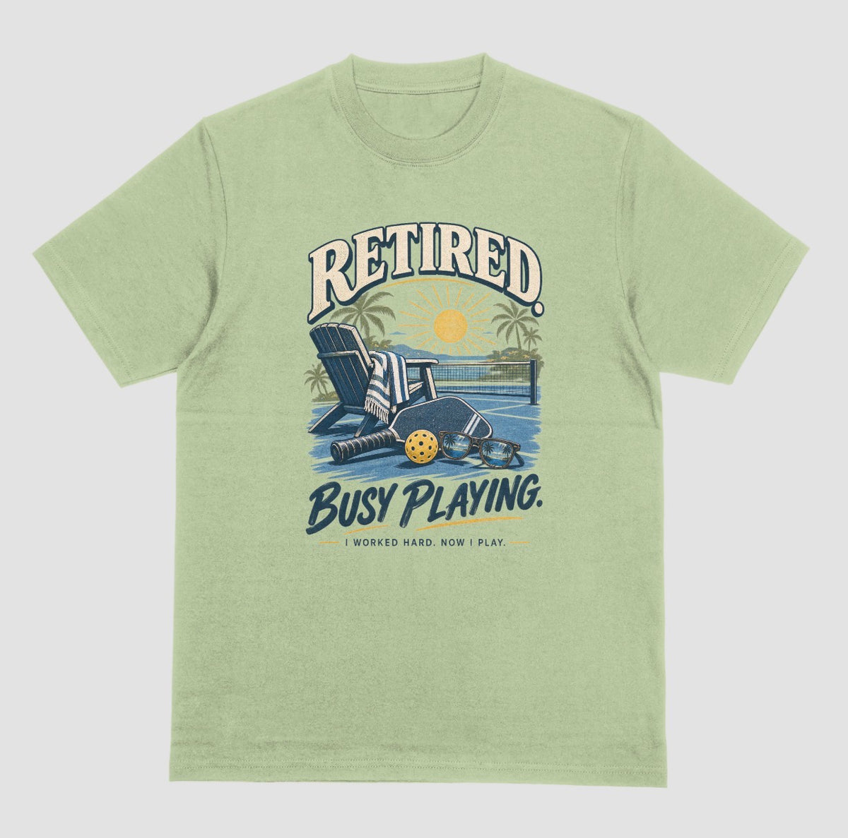 A light green short-sleeve t-shirt with a retro graphic on the chest. The graphic features the words Retired pickleball shirt in sand with a beach chair and paddle design that says Retired, Busy Playing, I Worked Hard Now I Play