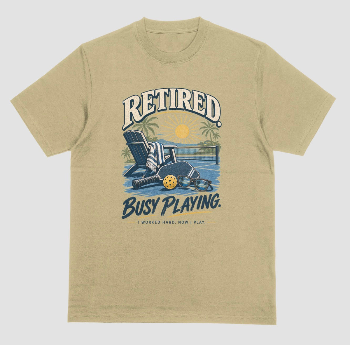 A sand color short-sleeve t-shirt with a retro graphic on the chest. The graphic features the words Retired pickleball shirt in sand with a beach chair and paddle design that says Retired, Busy Playing, I Worked Hard Now I Play