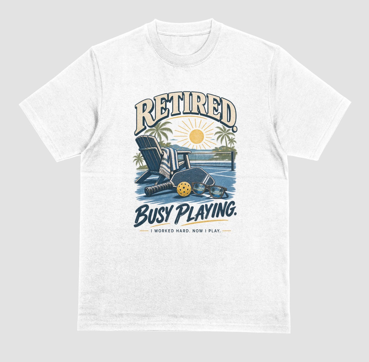 A white short-sleeve t-shirt with a retro graphic on the chest. The graphic features the words Retired pickleball shirt in sand with a beach chair and paddle design that says Retired, Busy Playing, I Worked Hard Now I Play