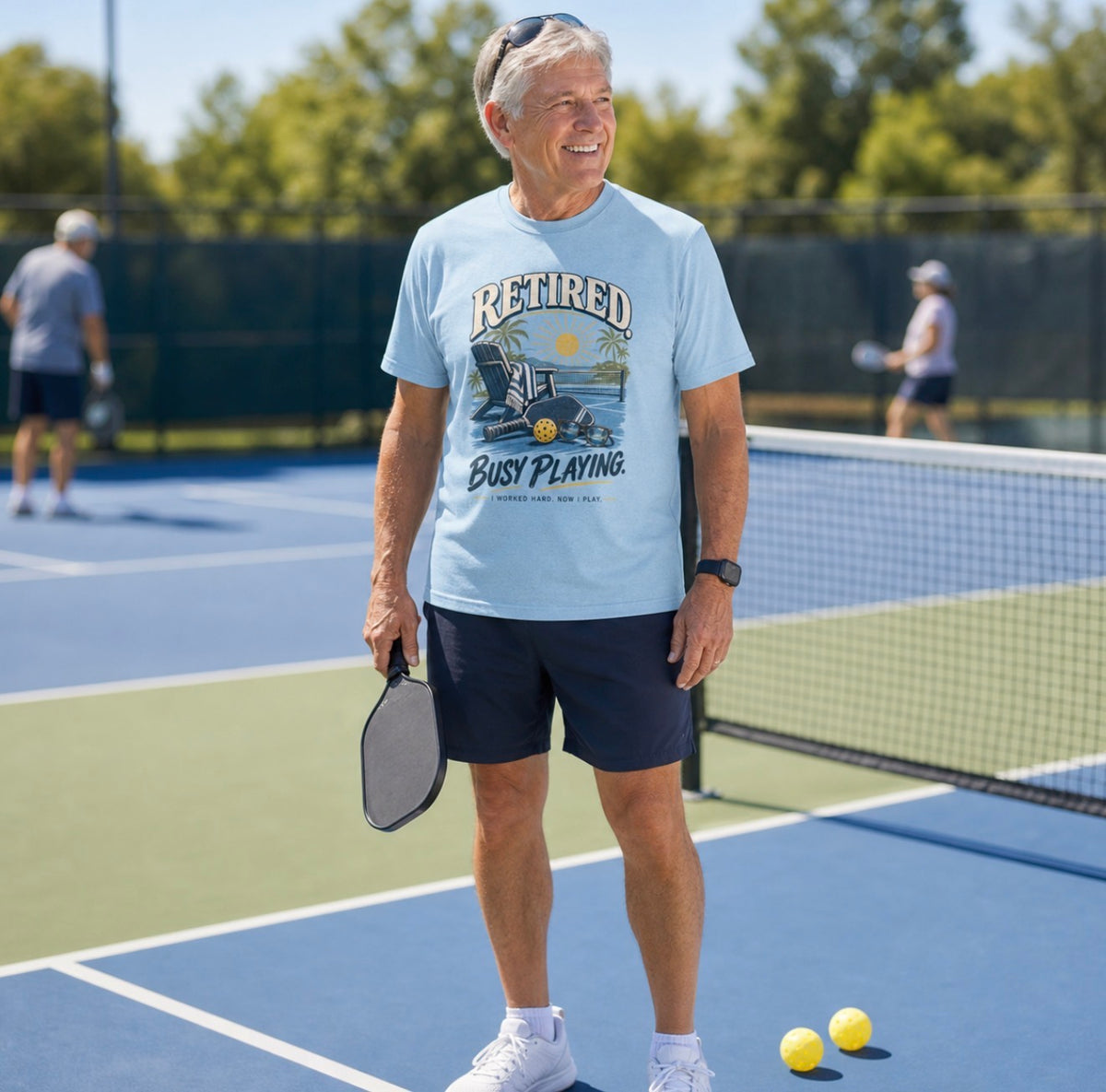 A man on a Pickleball court wearing short-sleeve t-shirt with a retro graphic on the chest. The graphic features the words short-sleeve t-shirt with a retro graphic on the chest. The graphic features the words Retired pickleball shirt in sand with a beach chair and paddle design that says Retired, Busy Playing, I Worked Hard Now I Play