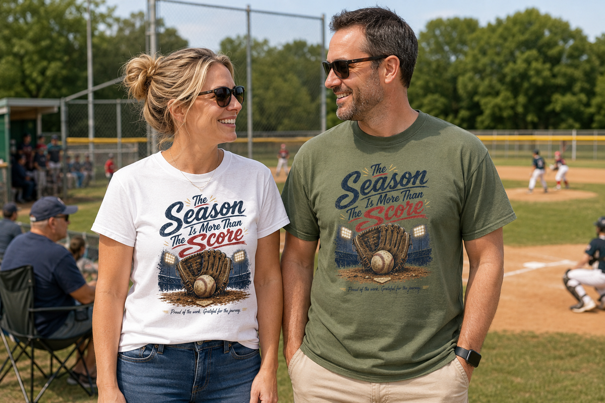 A man and a woman standing on a baseball field wearing matching graphic t-shirts featuring a vintage-style baseball graphic. The design reads “The Season Is More Than The Score” in navy blue and red lettering above a brown leather baseball glove holding a baseball, with a baseball field and illuminated stadium lights in the background. The design also includes the phrase “Proud of the work. Grateful for the journey.”