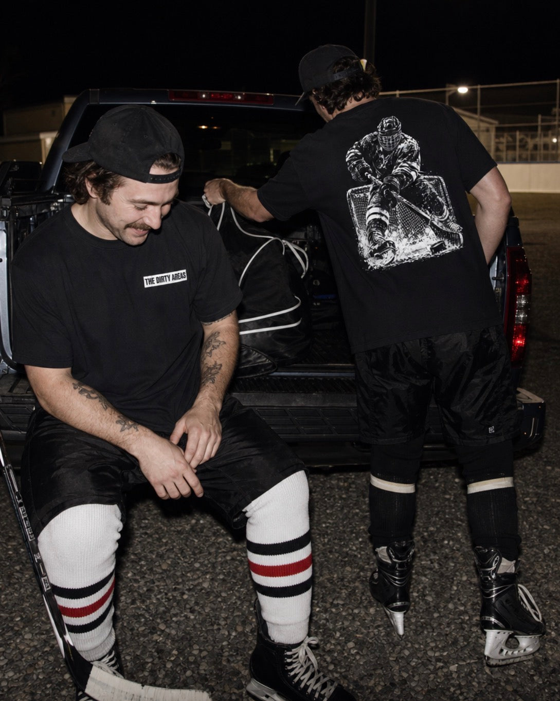 Two hockey players at a night tailgate in black 'The Dirty Areas' tees: small label on the chest, large hockey player in front of the net on the back.