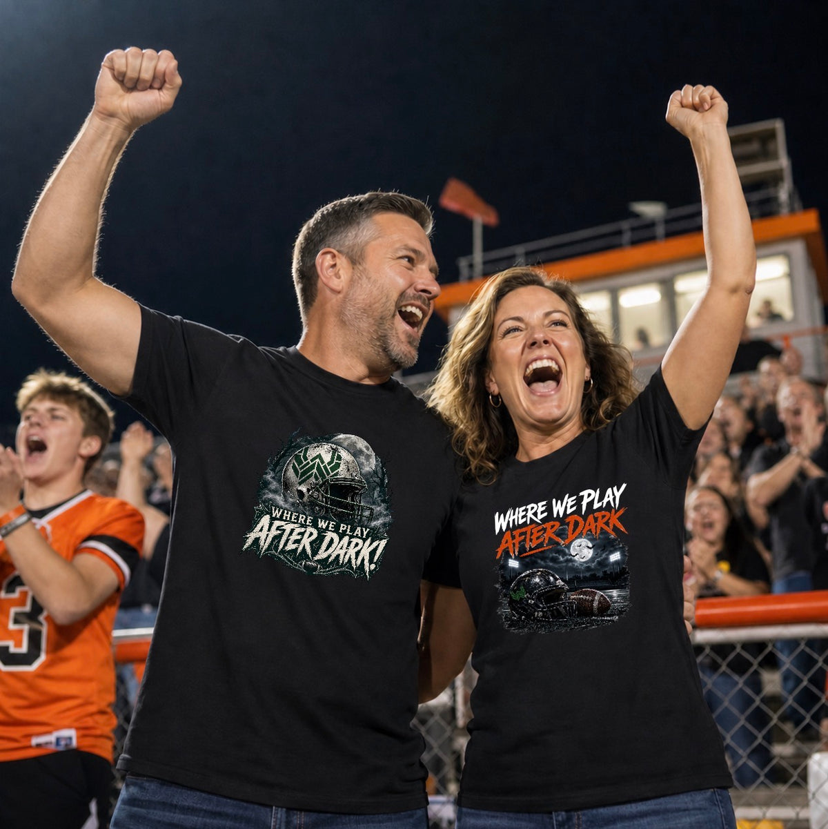 "At a Friday night football game, a smiling man and woman stand arm in arm, each with one fist raised and mouths open in a cheer. They both wear black t-shirts from the 'Where We Play After Dark' collection: his has the green-and-silver helmet and moon design, and hers has the orange-and-white lettering over a helmet and football on a night field. Behind them, an orange-trimmed press box and a crowd of fans, including a teen in an orange jersey, fill the stands under stadium lights."