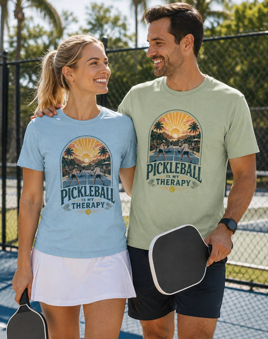 A man and a woman standing on a pickleball court wearing matching graphic t-shirts. The woman's shirt is light blue and the man's shirt is sage green, both featuring a retro sunset beach scene with the text 'PICKLEBALL IS MY THERAPY'.