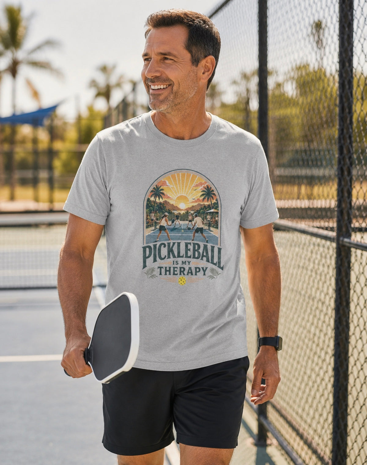 A man on a pickleball court wearing a light gray crewneck t-shirt featuring a retro-style graphic of pickleball players on a court with a sunset and palm trees, and the text 'PICKLEBALL IS MY THERAPY' printed below.