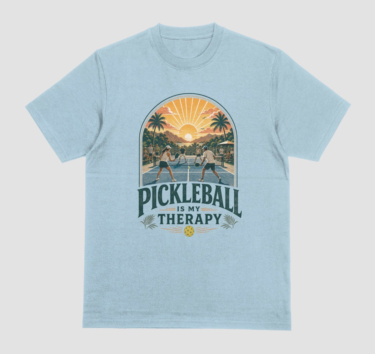 A light blue crewneck t-shirt featuring a retro-style graphic of pickleball players on a court with a sunset and palm trees, and the text 'PICKLEBALL IS MY THERAPY' printed below.