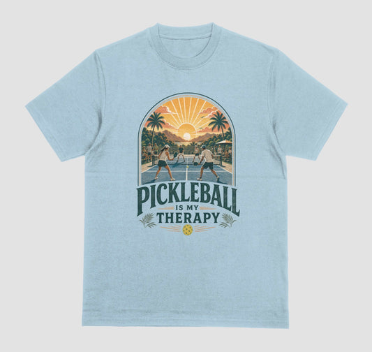 A light blue crewneck t-shirt featuring a retro-style graphic of pickleball players on a court with a sunset and palm trees, and the text 'PICKLEBALL IS MY THERAPY' printed below.