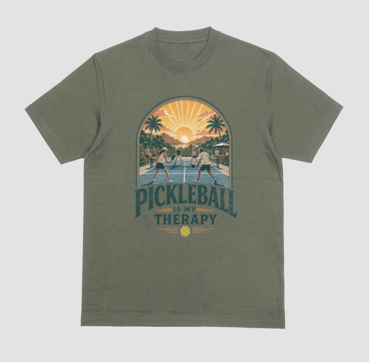 A military green crewneck t-shirt featuring a retro-style graphic of pickleball players on a court with a sunset and palm trees, and the text 'PICKLEBALL IS MY THERAPY' printed below.