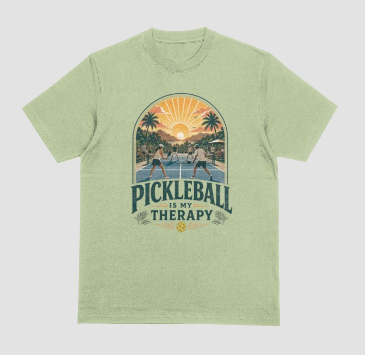 A pistachio crewneck t-shirt featuring a retro-style graphic of pickleball players on a court with a sunset and palm trees, and the text 'PICKLEBALL IS MY THERAPY' printed below.