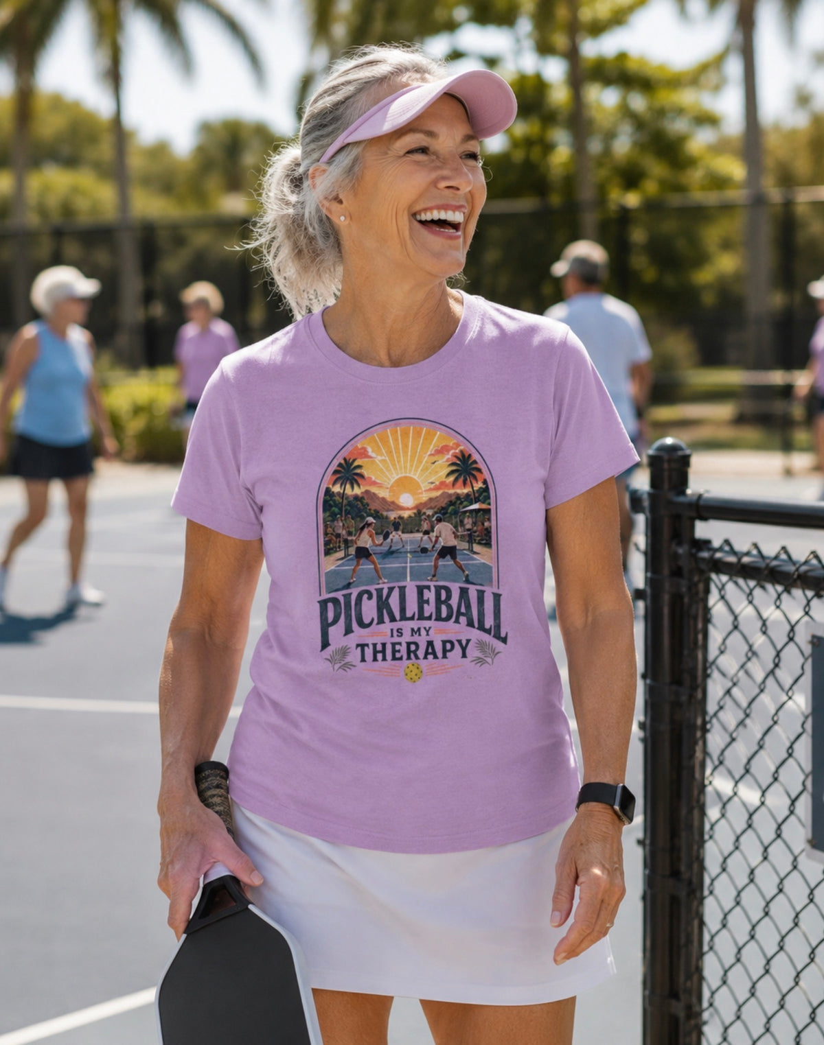 A woman on an outdoor pickleball court wearing a lilac graphic t-shirt that reads 'PICKLEBALL IS MY THERAPY' with a retro pickleball court scene, paired with a white skirt and pink visor.